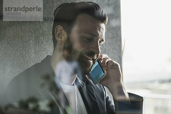 Lächelnder Geschäftsmann  der im Büro durch Glas gesehen am Smartphone spricht