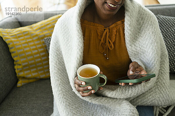 Lächelnde Frau  in eine Decke gehüllt  benutzt Smartphone und hält eine Tasse Tee in der Hand