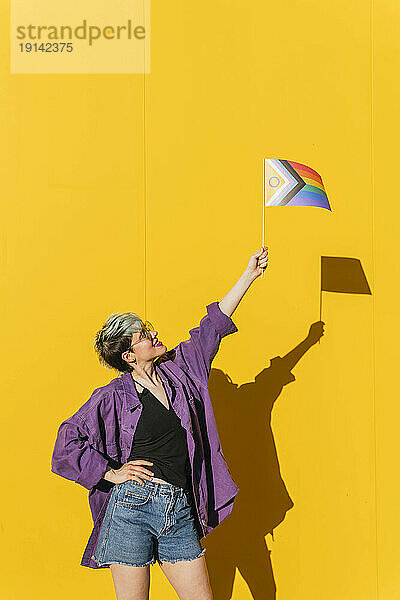 Lesbische Frau mit Hand an der Hüfte und Regenbogenfahne vor gelber Wand