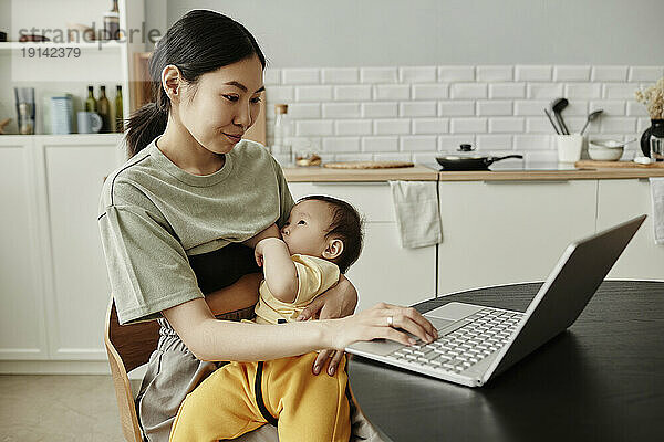 Smiling freelancer breastfeeding daughter and using laptop at home
