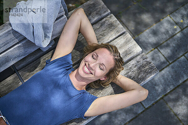 Carefree smiling woman with hands behind head lying on bench at sunny day