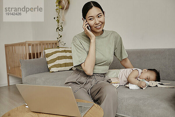 Freelancer mother talking on smart phone with daughter sleeping on sofa at home