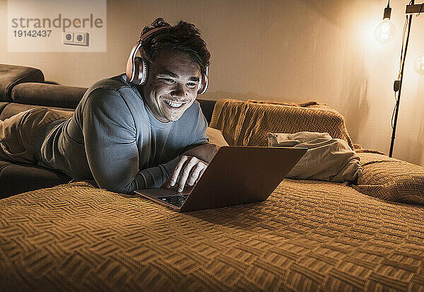 Junger Mann trägt kabellose Kopfhörer und benutzt Laptop im Wohnzimmer