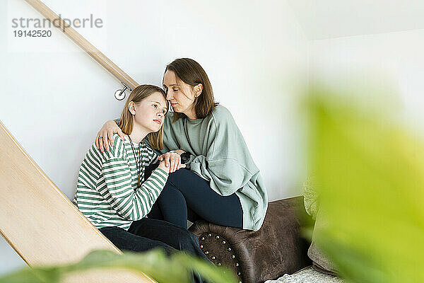 Mother consoling sad daughter sitting near staircase at home
