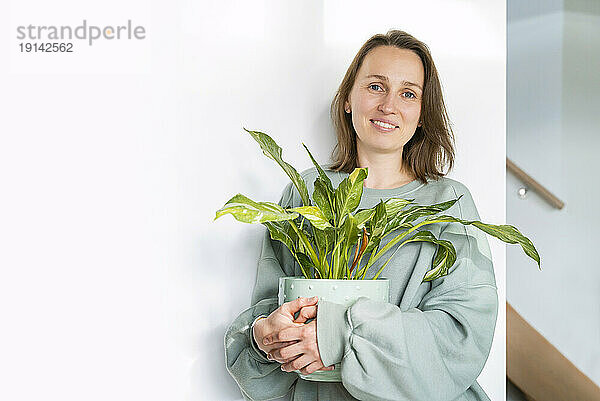 Lächelnde Frau hält zu Hause eine Topfpflanze an der Wand