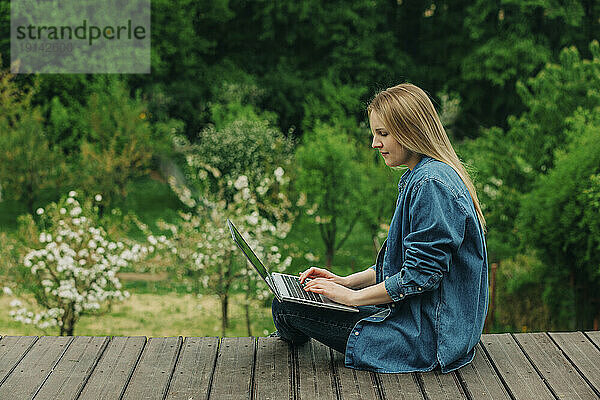 Blonde Frau sitzt mit Laptop auf Holzsteg im Garten