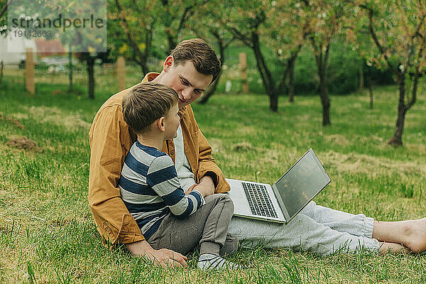 Sohn und Vater sitzen mit Laptop im Gras im Garten