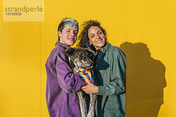 Smiling lesbian couple carrying dog in front of yellow wall