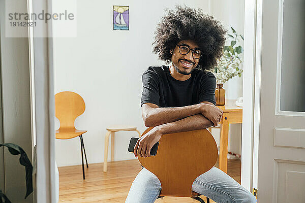 Lächelnder Mann mit Smartphone sitzt zu Hause auf einem Stuhl