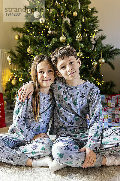 Smiling brother and sister sitting in front of Christmas tree