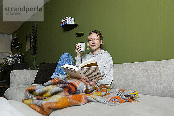 Junge Frau mit Tasse liest Buch auf Couch