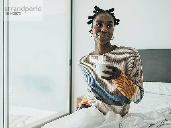 Glückliche junge Frau  die zu Hause auf dem Bett sitzt und eine Tasse Kaffee trinkt