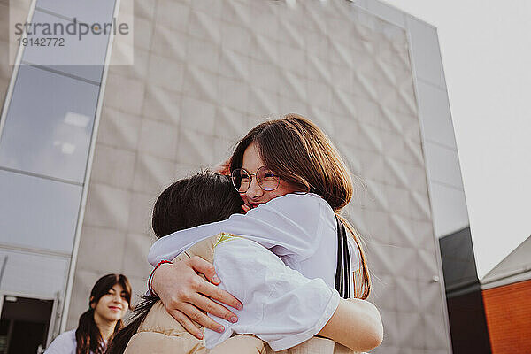 Happy girl embracing friend standing in front of school building