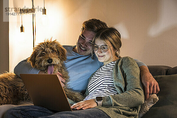 Smiling couple using laptop and sitting with dog on sofa at home
