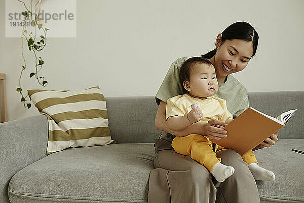 Happy mother reading book with daughter sitting on sofa at home