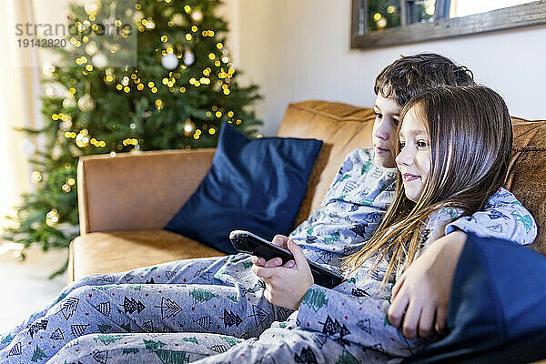 Lächelndes Mädchen  das zur Weihnachtszeit zu Hause mit seinem Bruder fernsieht