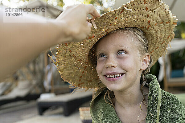 Frau setzt lächelndem Mädchen am Strand Strohhut auf