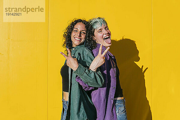 Fröhliche lesbische Frauen zeigen Friedenszeichen vor gelber Wand