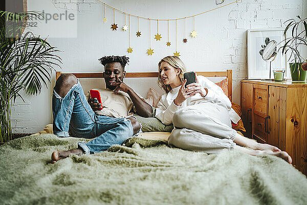 Junge Frau schaut auf das Smartphone ihres Freundes und entspannt sich im Bett