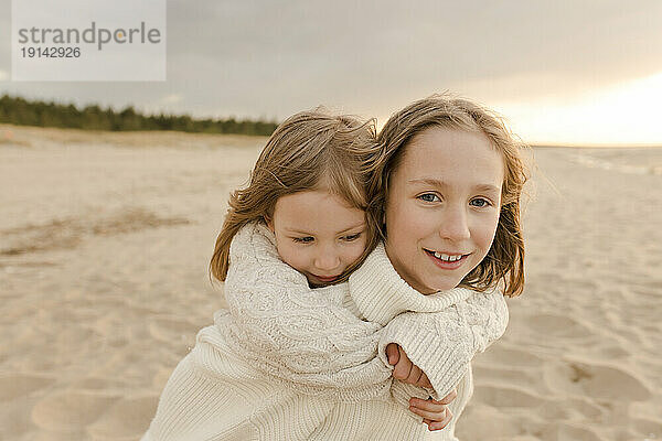 Glückliches Mädchen  das seiner Schwester Huckepackfahrt am Strand gibt