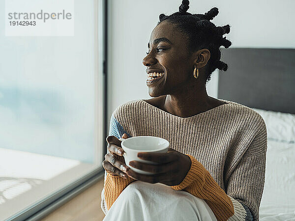 Lächelnde Frau sitzt auf dem Bett und hält eine Kaffeetasse in der Hand