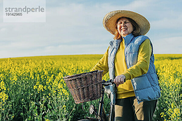 Lächelnde ältere Frau mit Fahrrad steht im Rapsfeld