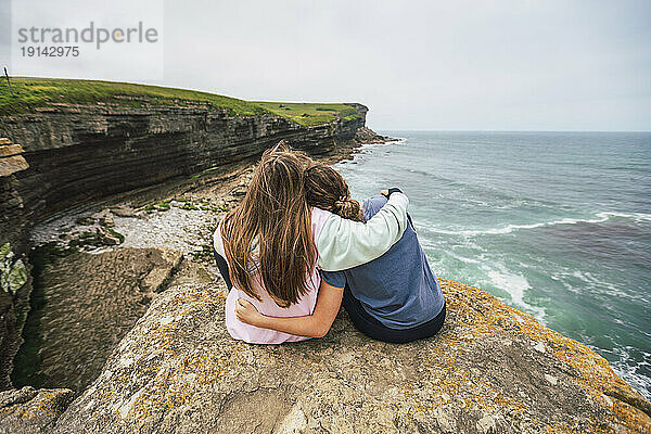 Frau mit Arm um Freundin sitzt auf Felsen und blickt aufs Meer