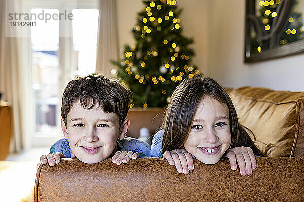Glückliches Mädchen mit Bruder  der sich zur Weihnachtszeit zu Hause auf dem Sofa amüsiert