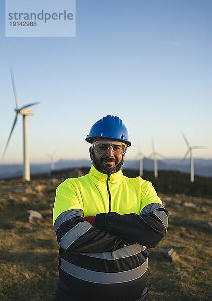 Lächelnder Ingenieur mit verschränkten Armen steht vor Windkraftanlagen