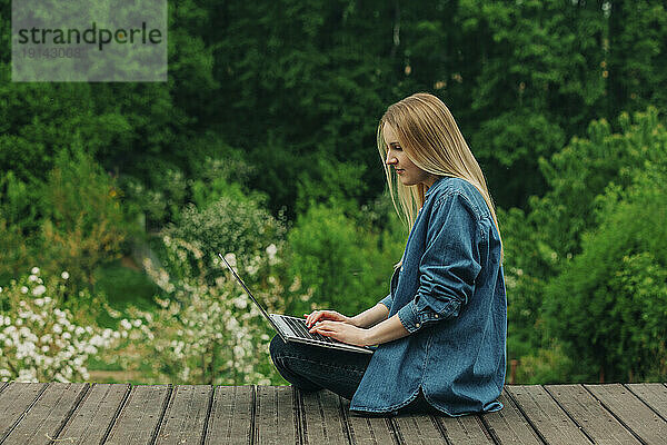 Frau sitzt mit Laptop auf Holzsteg im Garten