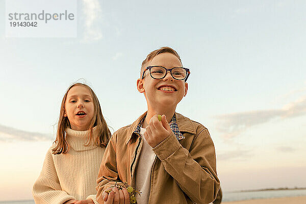 Glücklicher Junge mit Schwester  die unter dem Himmel am Strand steht