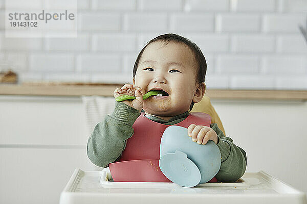 Smiling girl holding spoon in mouth sitting on high chair at home