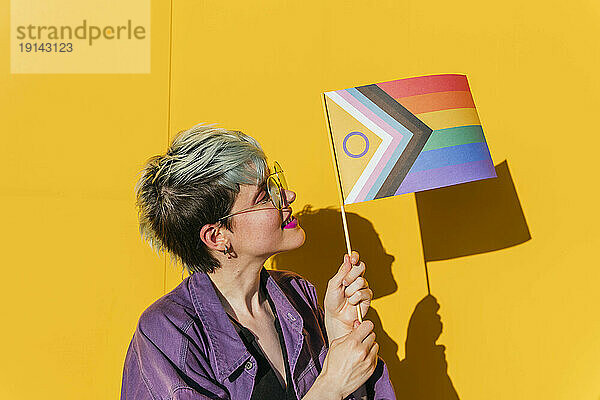 Lesbische Frau mit gefärbten Haaren hält Regenbogenfahne vor gelber Wand