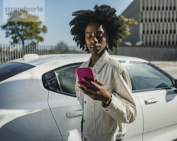 Junge Frau benutzt Smartphone und steht neben weißem Auto