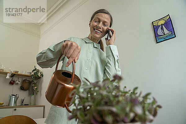 Glückliche Frau  die zu Hause auf dem Smartphone spricht und Pflanzen gießt