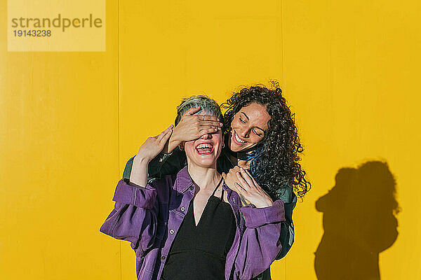 Glückliche Frau bedeckt die Augen einer lesbischen Freundin vor einer gelben Wand