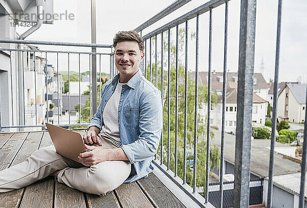 Glücklicher Mann sitzt mit Laptop auf dem Balkon