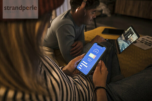 Junge Frau bedient Elektroauto-Lade-App auf Smartphone und Mann benutzt Laptop im Hintergrund