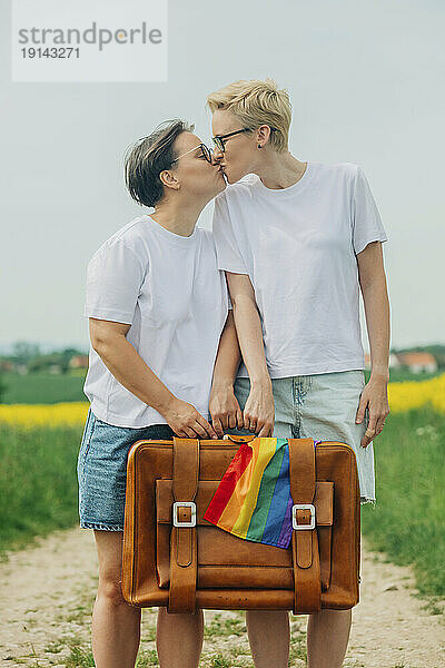 Liebevolle Lesben halten Koffer mit Regenbogenfahne in der Hand und küssen sich