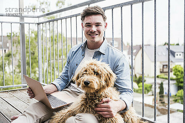 Glücklicher junger Mann sitzt mit Hund und Laptop auf dem Balkon
