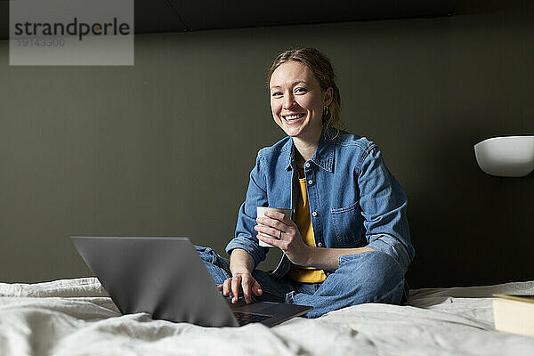 Lächelnde Geschäftsfrau hält Kaffeetasse mit Laptop auf dem Bett