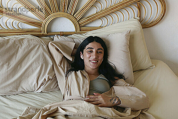 Lächelnde junge Frau entspannt sich im Bett und genießt zu Hause Musik