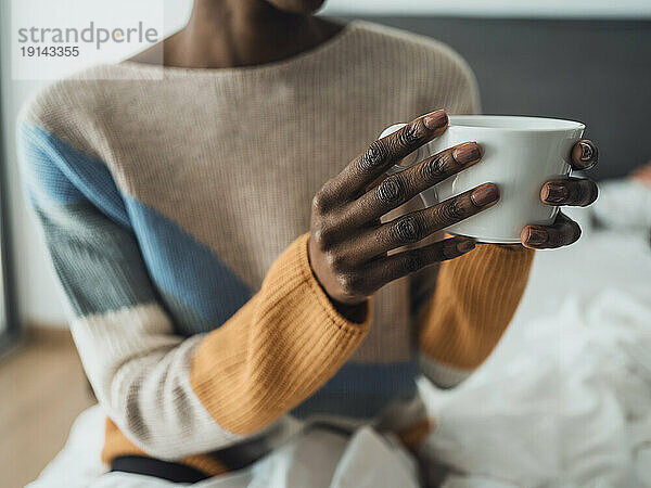 Hände einer Frau  die auf dem Bett sitzt und eine Tasse Kaffee hält