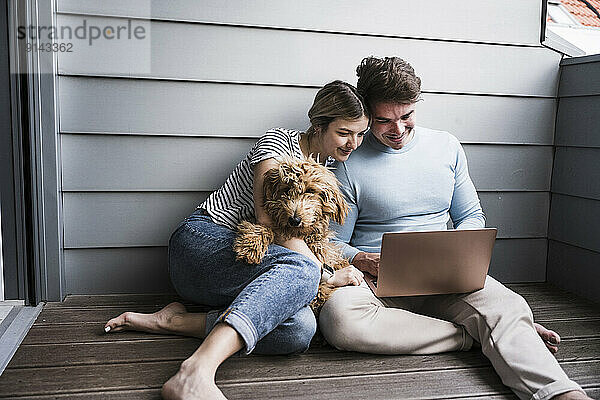 Smiling man and woman using laptop and sitting with dog in front of wall