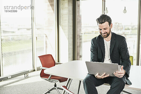Lächelnder Geschäftsmann sitzt mit Laptop am Tisch im Büro