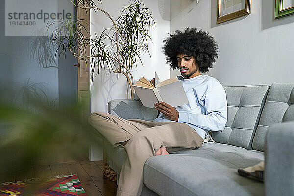 Mann liest ein Buch und sitzt zu Hause auf dem Sofa