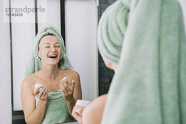 Glückliche Frau hält eine Flasche Gesichtscreme in der Hand und schaut in den Badezimmerspiegel