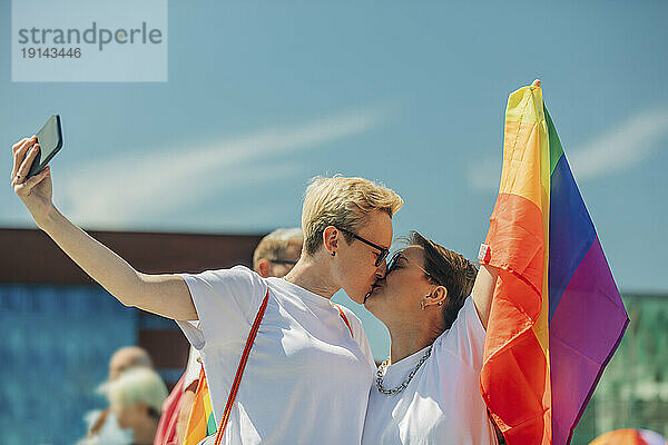 Lesbian couple kissing and taking selfie with rainbow flag