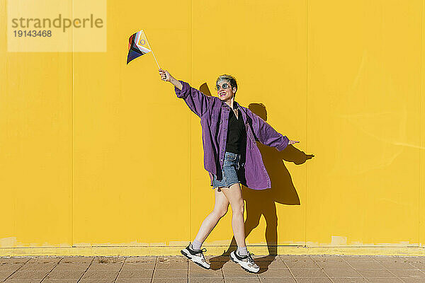 Sorglose lesbische Frau springt mit Regenbogenfahne vor gelber Wand