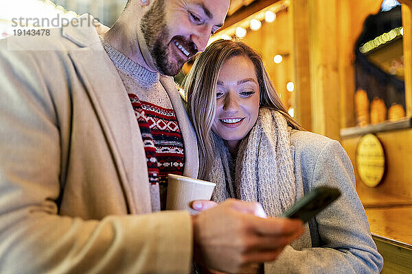 Lächelnde Frau und Mann benutzen Mobiltelefon auf dem Weihnachtsmarkt
